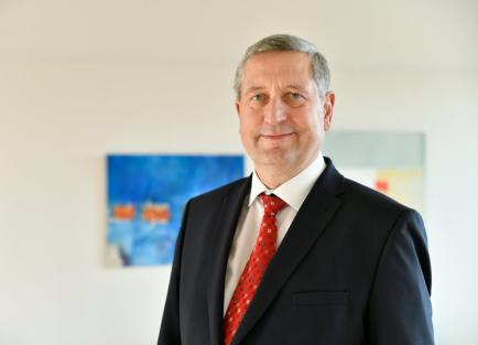 Mann mit grauen Haaren und blauen Augen bis zur Brust sichtbar mit schwarzem Anzug und roter Krawatte schaut in die Kamera.