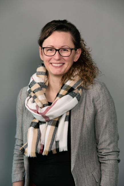 Dunkelhaarige Frau mit braunen Augen bis zum Bauch sichtbar mit schwarzem Oberteil und grauer Anzugjacke schaut in die Kamera.