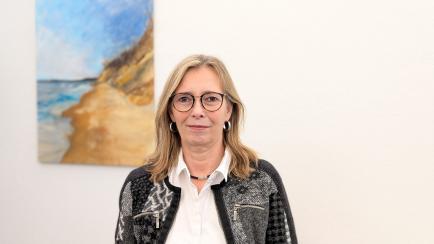 Blonde Frau mit braunen Augen in einer Porträtaufnahme mit weißem Oberteil und schwarz-grauer Jacke schaut in die Kamera.